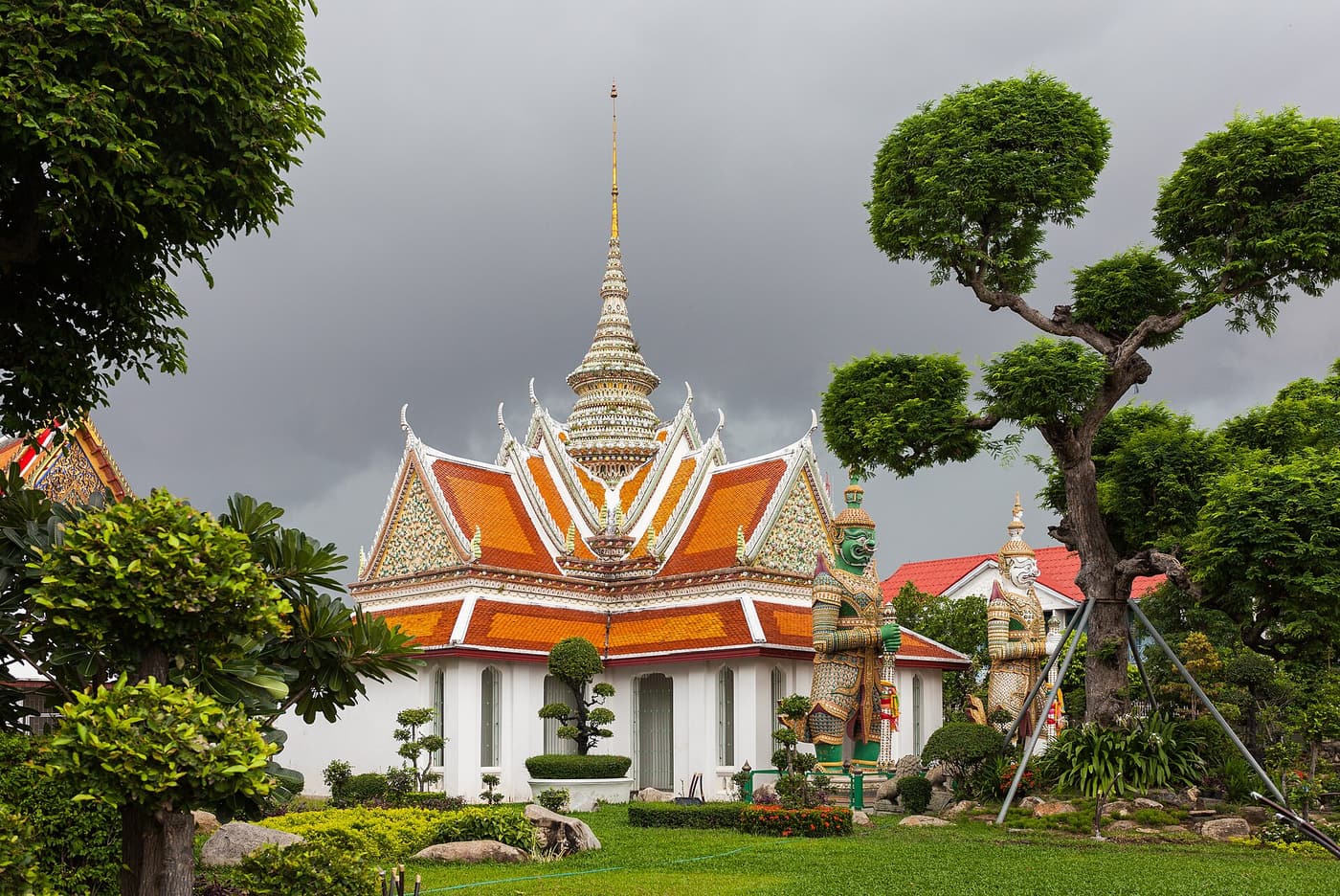 A white pavilion with orange-tiled roofs and a gilded spire in the grounds of Wat Arun in Bangkok, watched over by guardian statues.