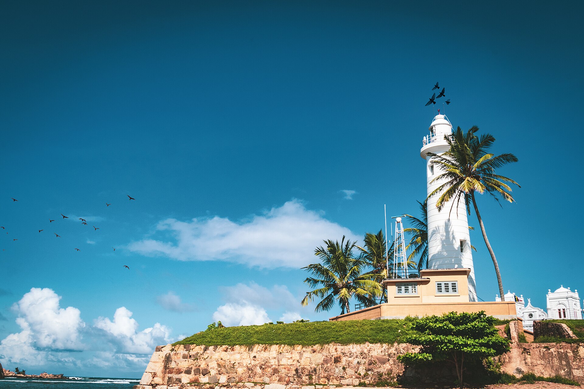 The white lighthouse and palm trees on the ramparts of Galle Fort, Sri Lanka, under a blue sky.
