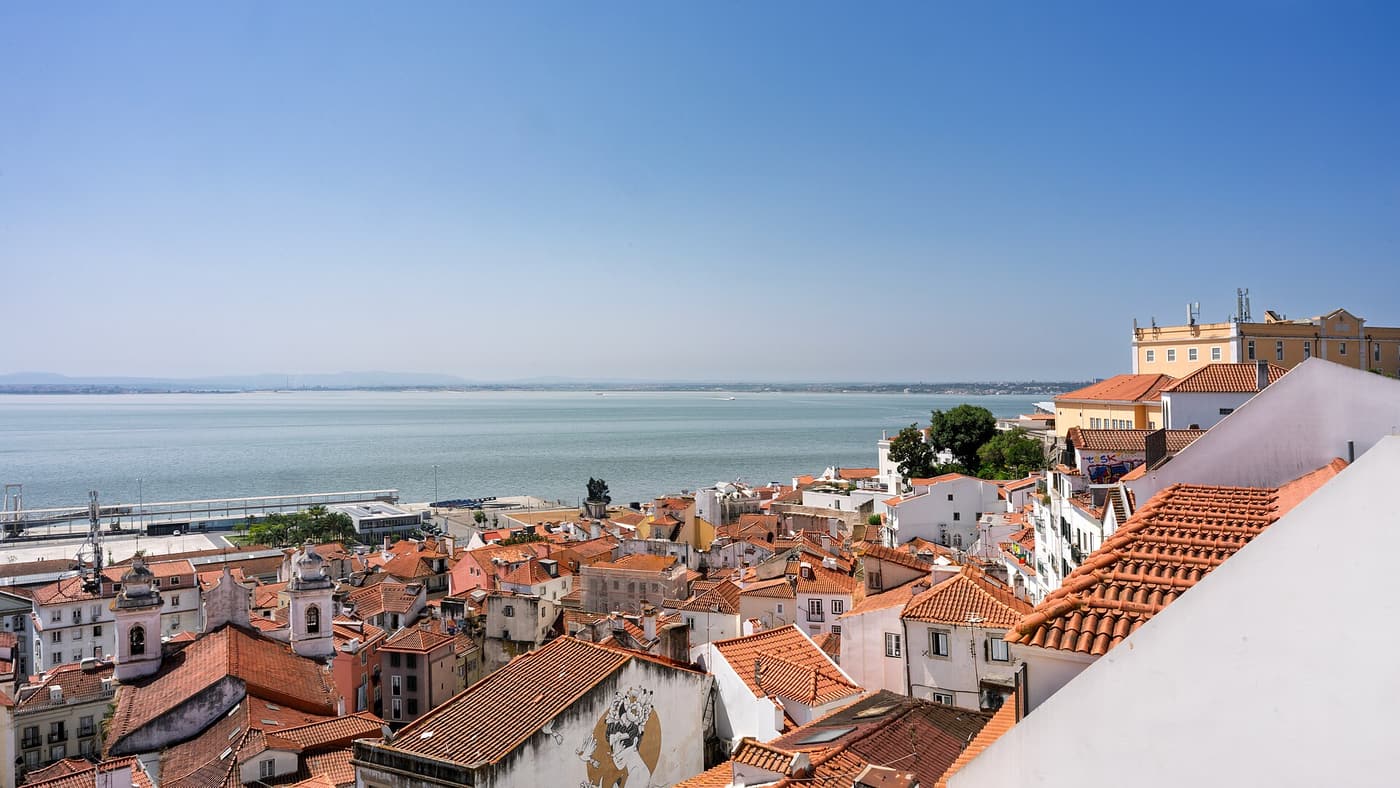 Terracotta rooftops of Alfama in Lisbon with the Tagus river beyond them.