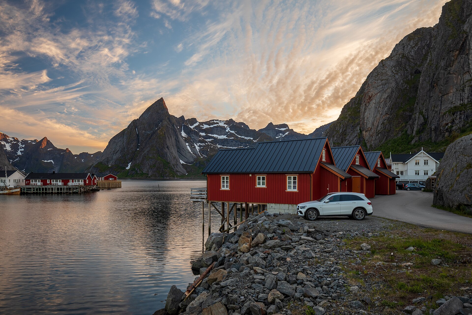 Red rorbu fishing cabins on stilts at the water's edge in Reine, Lofoten, below jagged peaks at dusk.