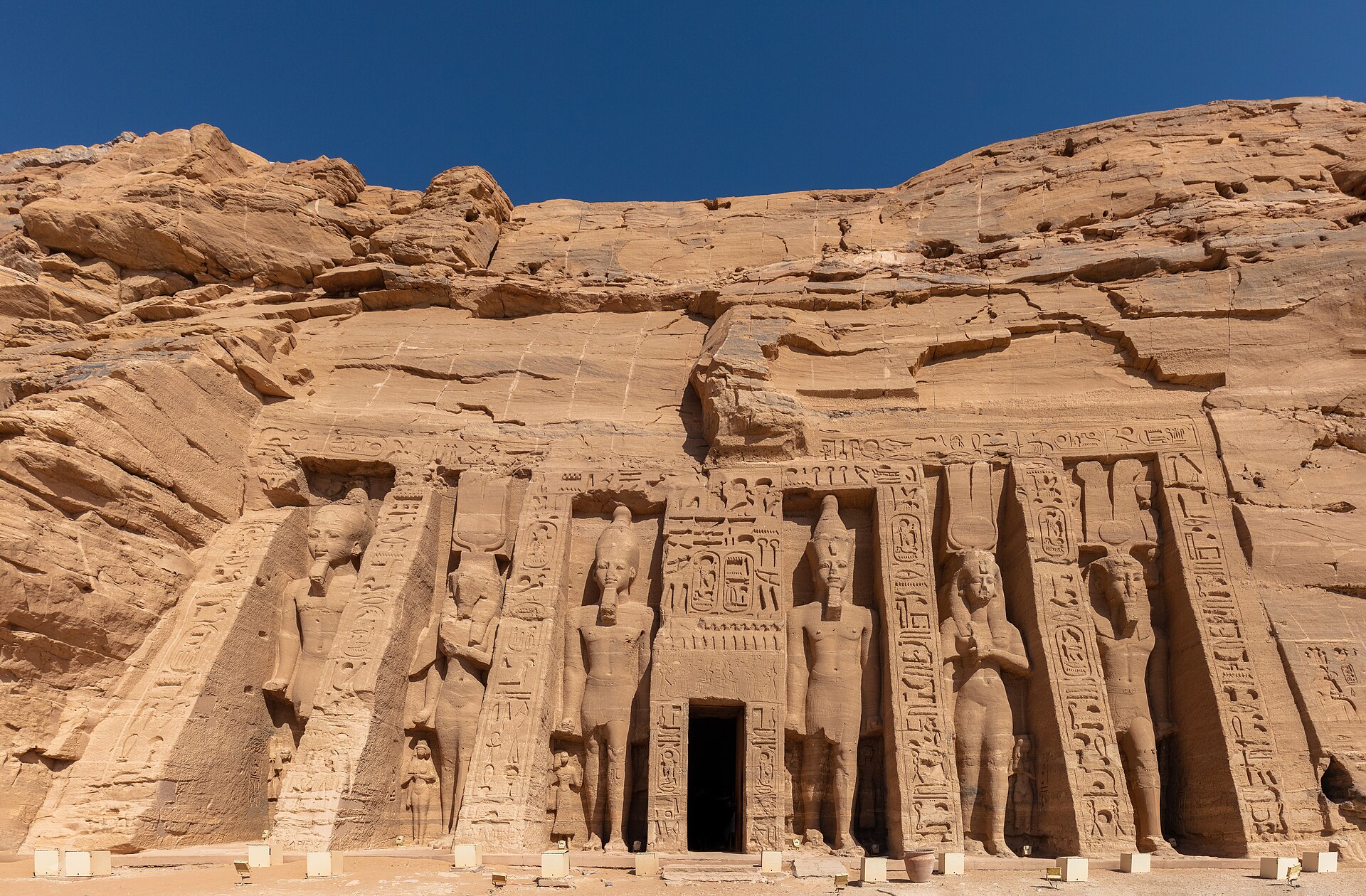 The rock-cut facade of the Temple of Nefertari at Abu Simbel, lined with standing statues under a clear sky.