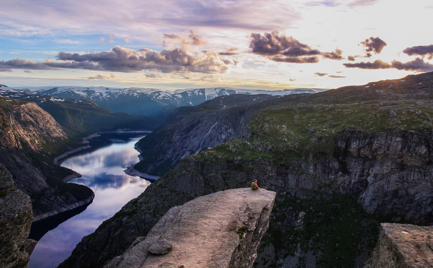 A lone hiker sitting at the end of the Trolltunga ledge above Ringedalsvatnet at sunset.
